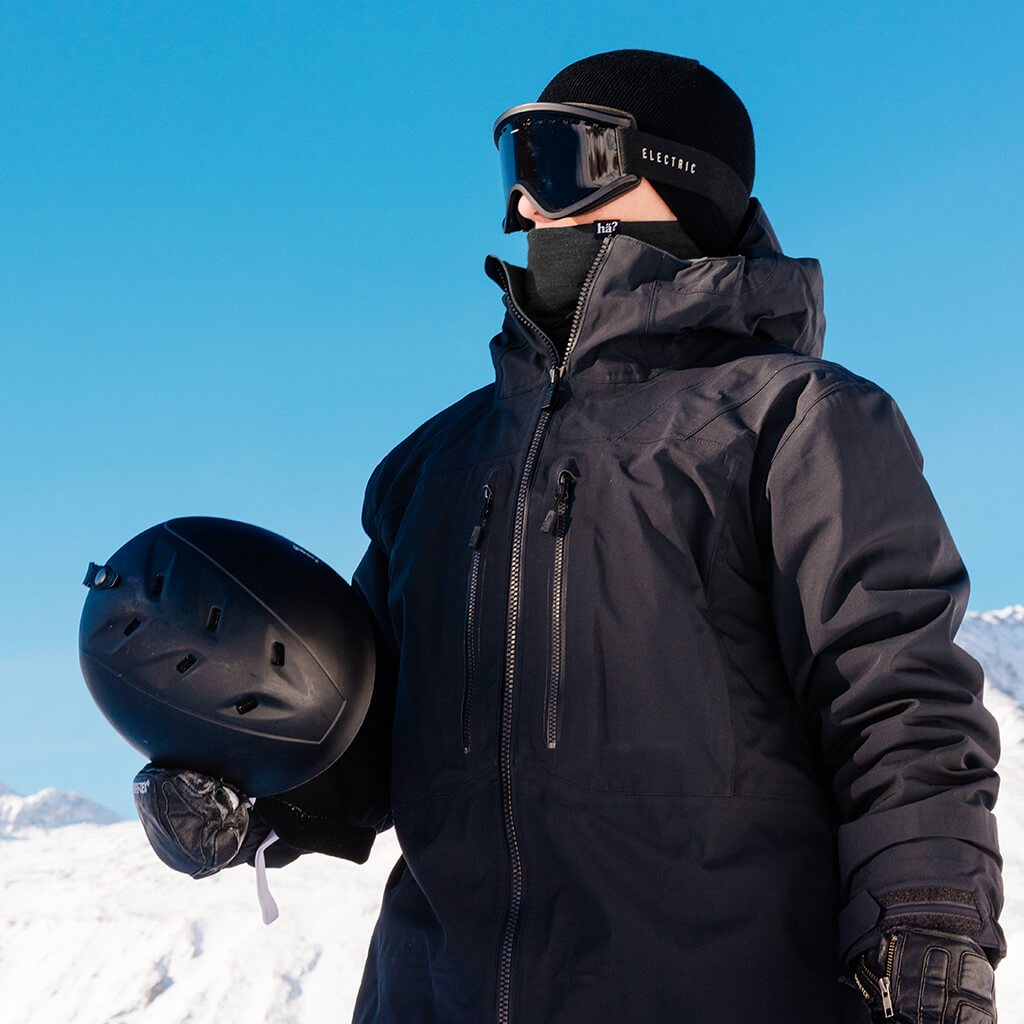 Ein Mann mit schwarzer Jacke, Beanie, Brille und Neckwarmer hält einen Helm in einer Winterlandschaft.