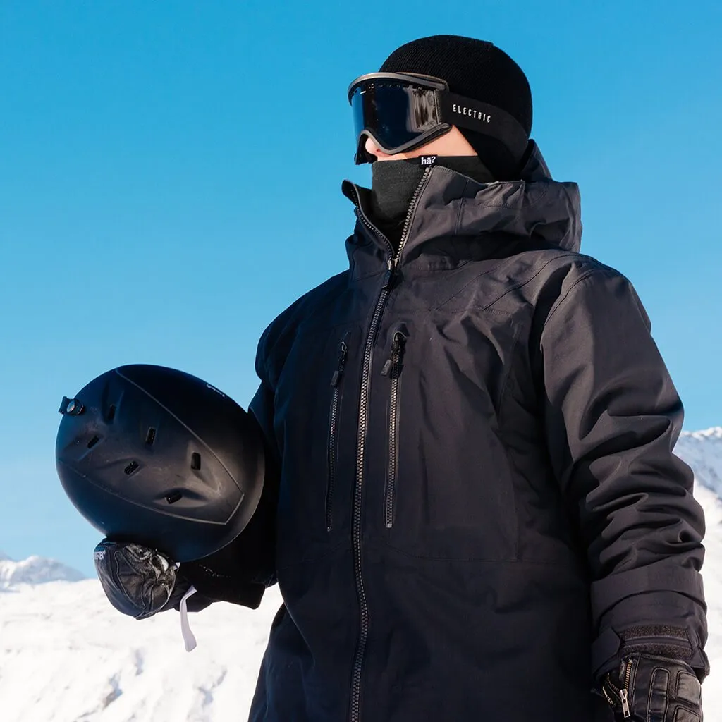 Un homme portant une veste noire, un bonnet, des lunettes et un cache-nez tient un casque dans un paysage hivernal.