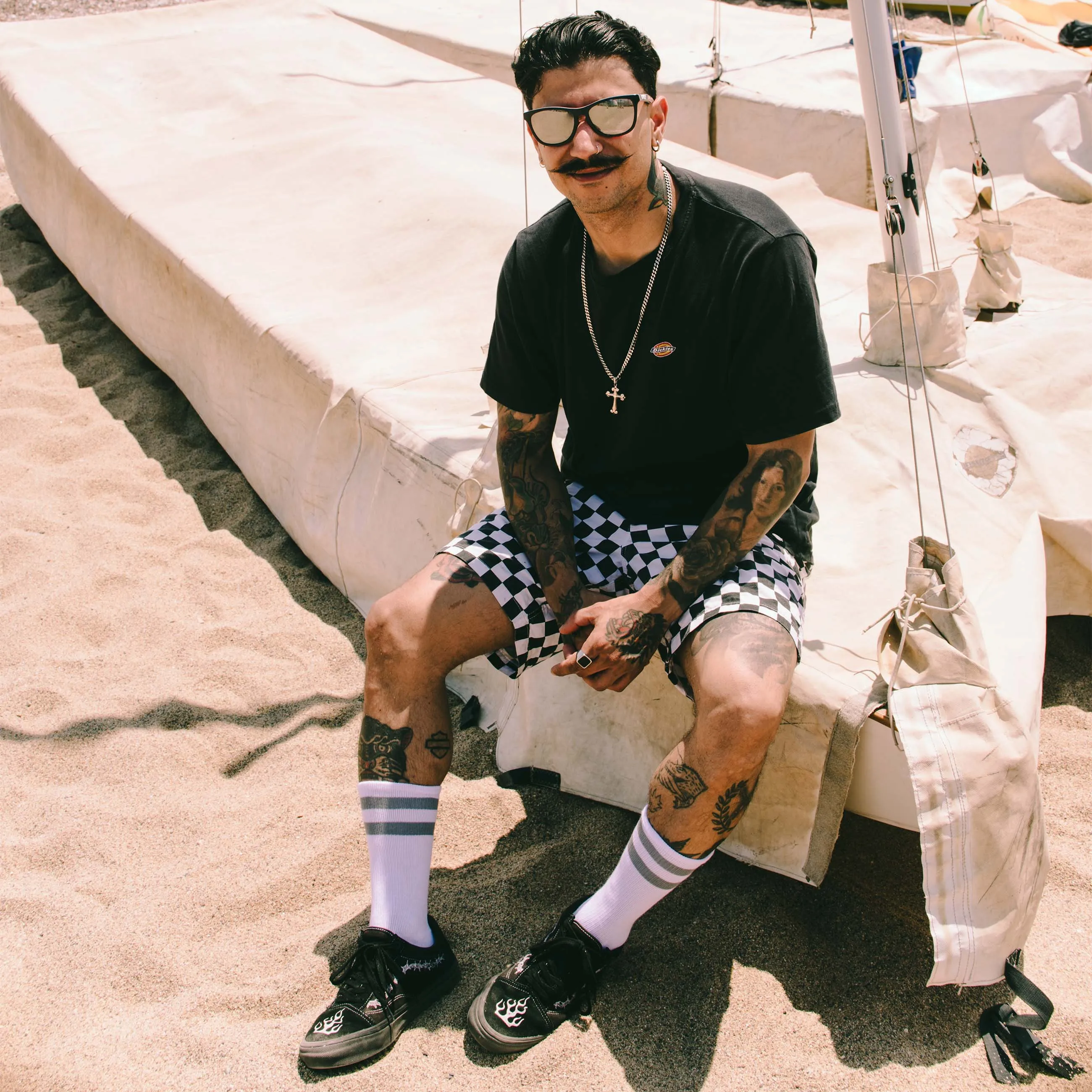 Mann mit Tätowierungen, Mid-High Socken und Schachbrettmuster-Shorts sitzt auf einer Plane am Strand.
