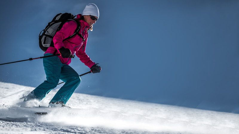 Une femme avec un sac à dos et des bâtons de ski dévale une pente enneigée avec des raquettes à neige Crossblades Softboot.