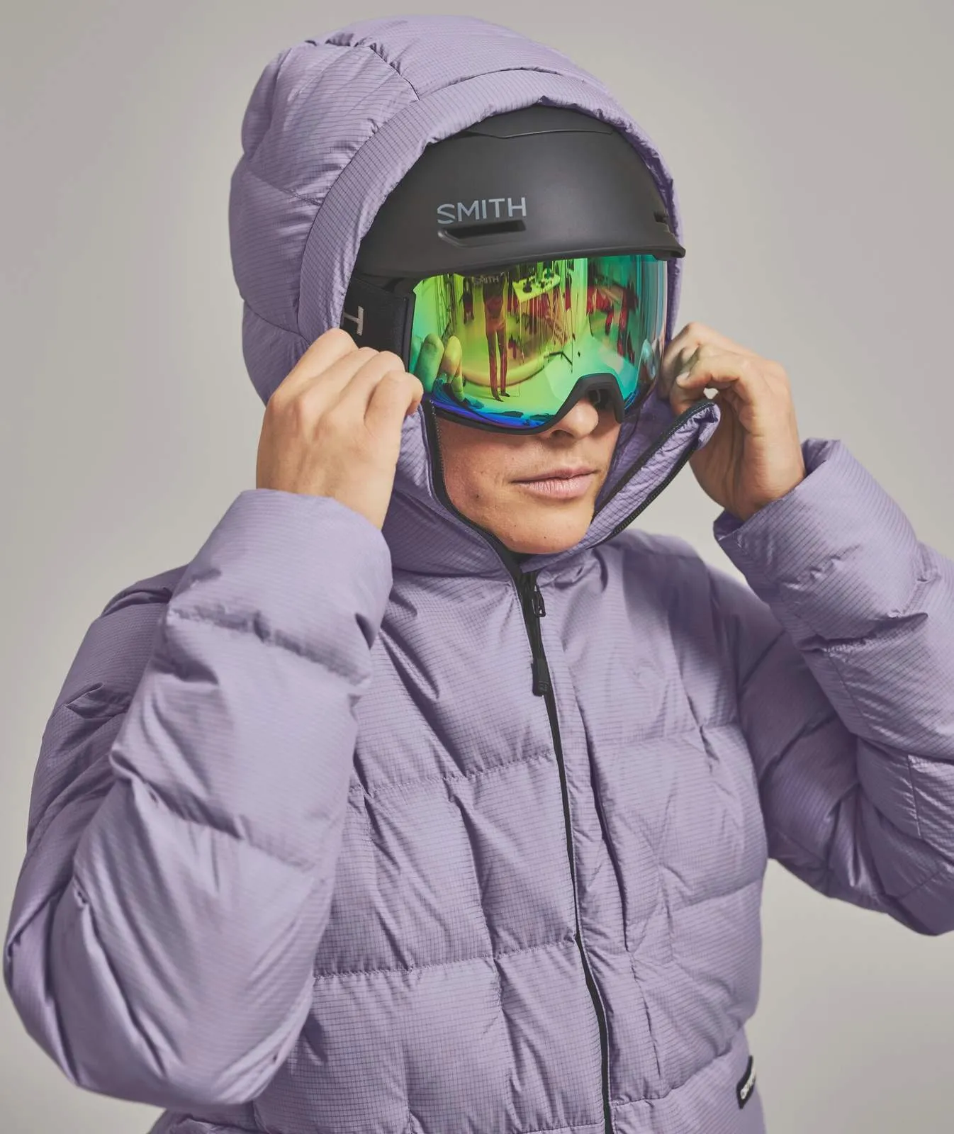 Person mit Skihelm und verspiegelter Brille unter Kapuze einer lila Daunenjacke. Reflexionen in der Brille.