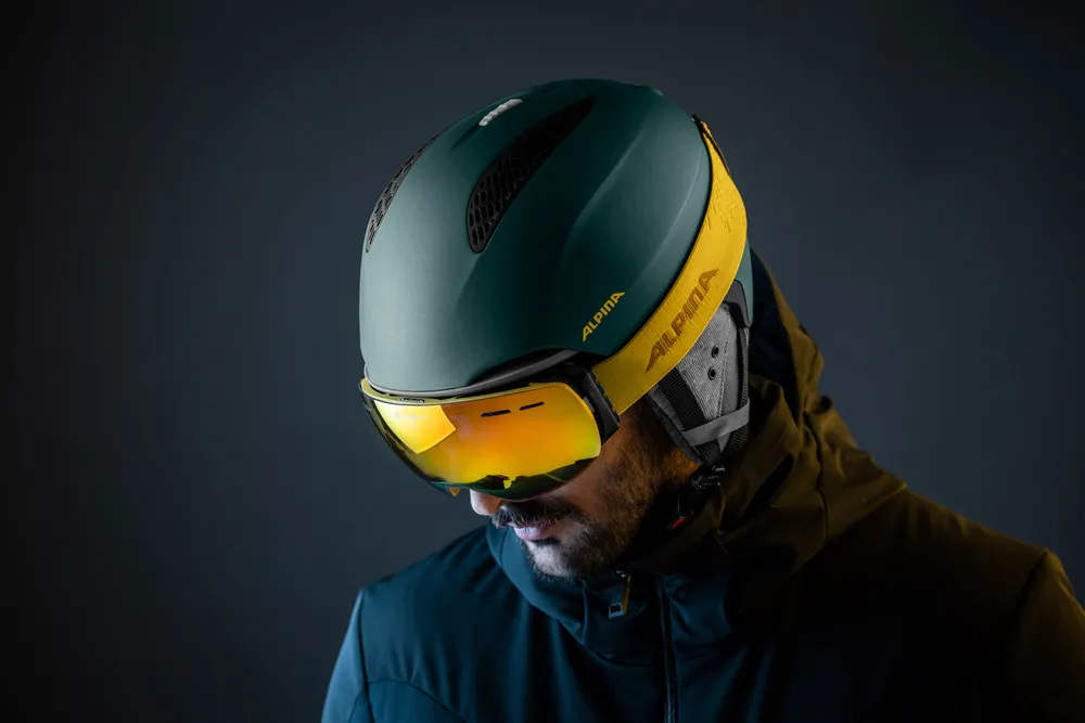 Uomo con casco da sci e occhiali gialli che guarda in basso; sfondo scuro.