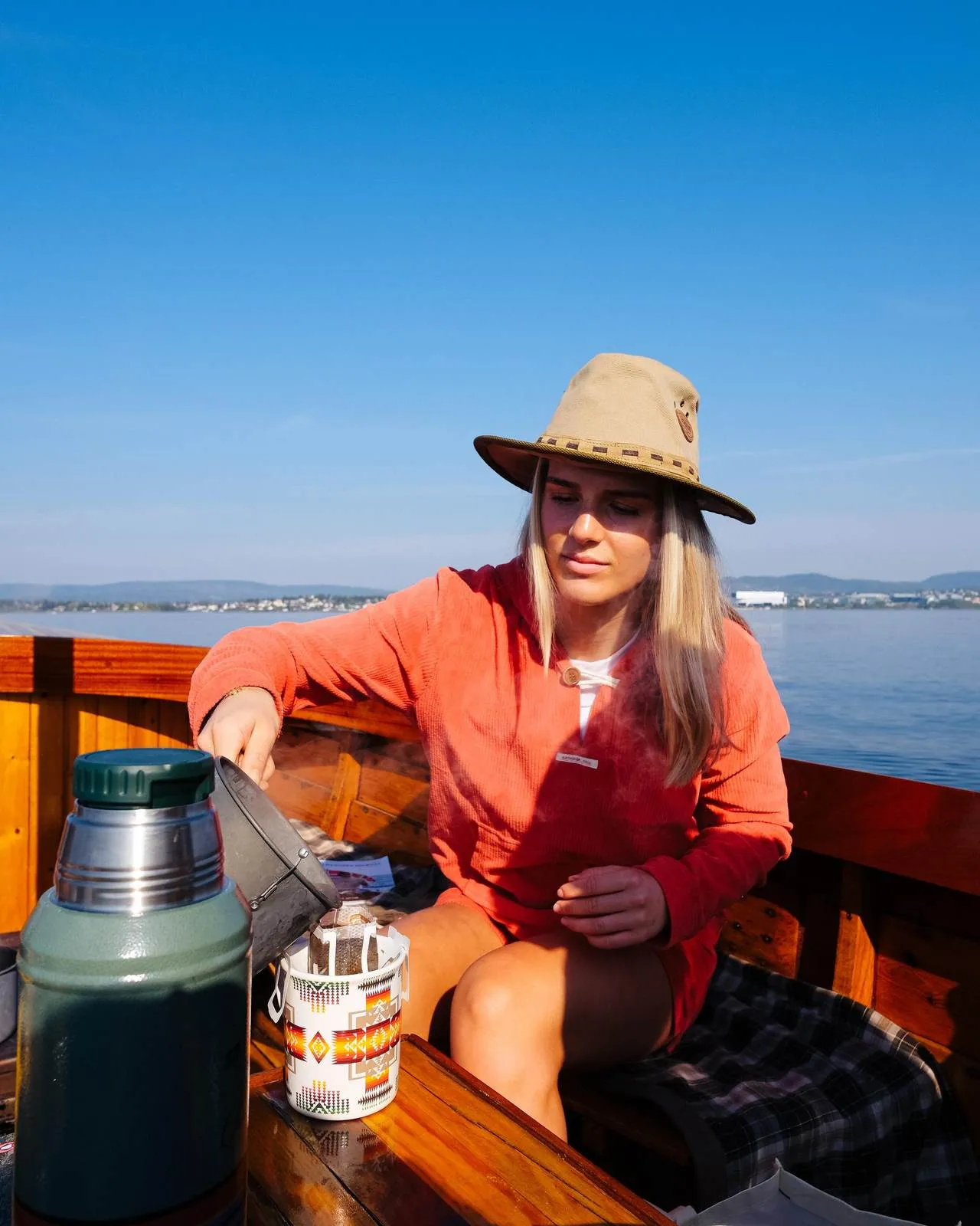 Una donna con un cappello versa del caffè fumante in una tazza su una barca di legno con il cielo blu e l'acqua sullo sfondo.