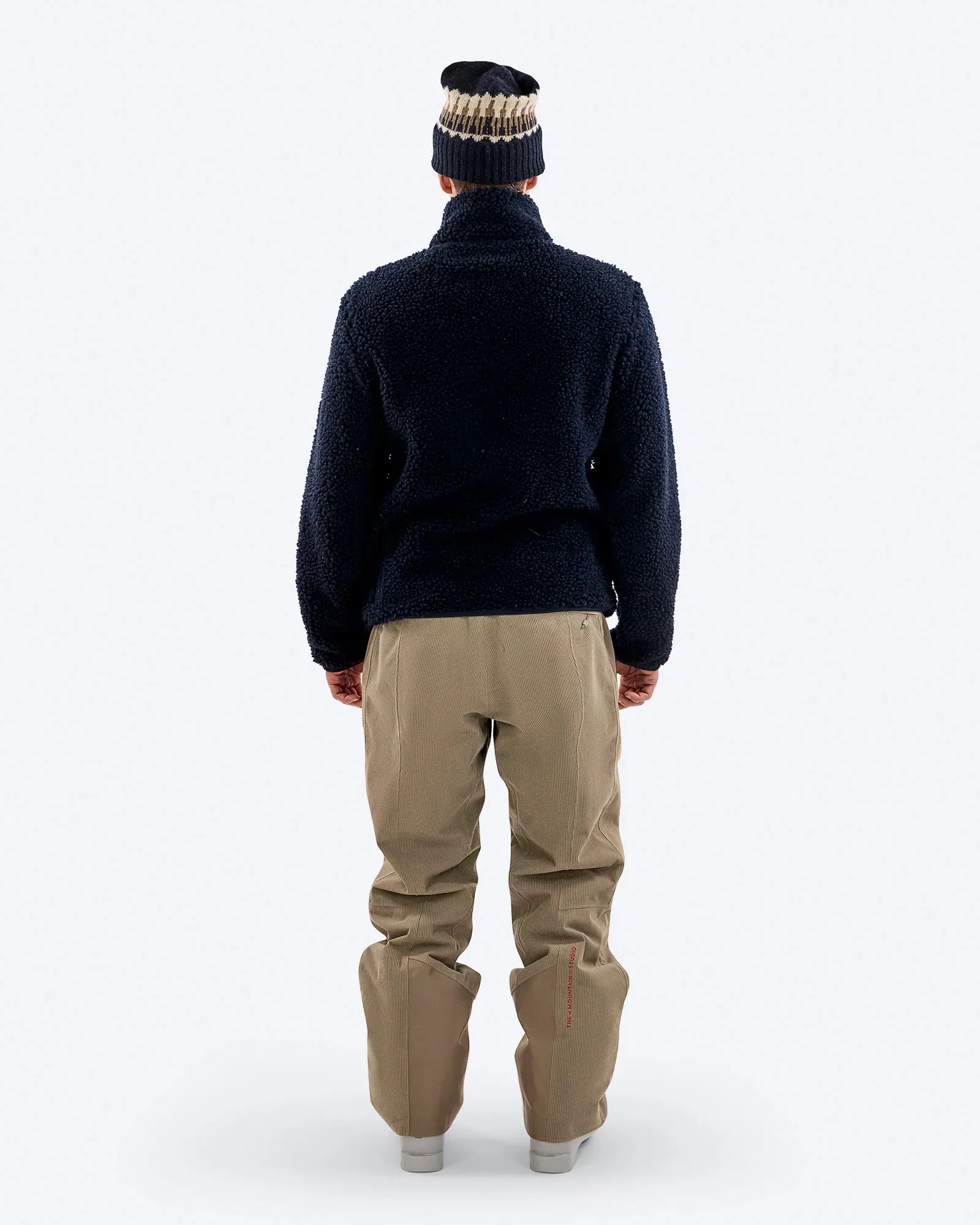 Persona di spalle che indossa un maglione di pile scuro, pantaloni beige e berretto, su sfondo bianco.