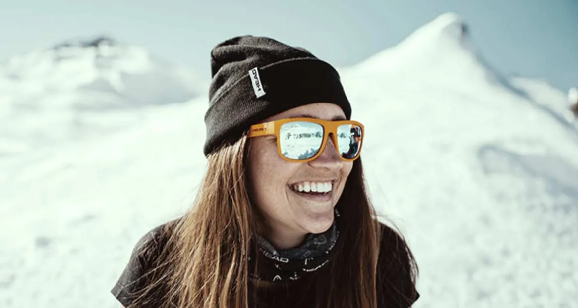 A woman smiles with orange sunglasses in front of snow-covered mountains. She is wearing a black cap.