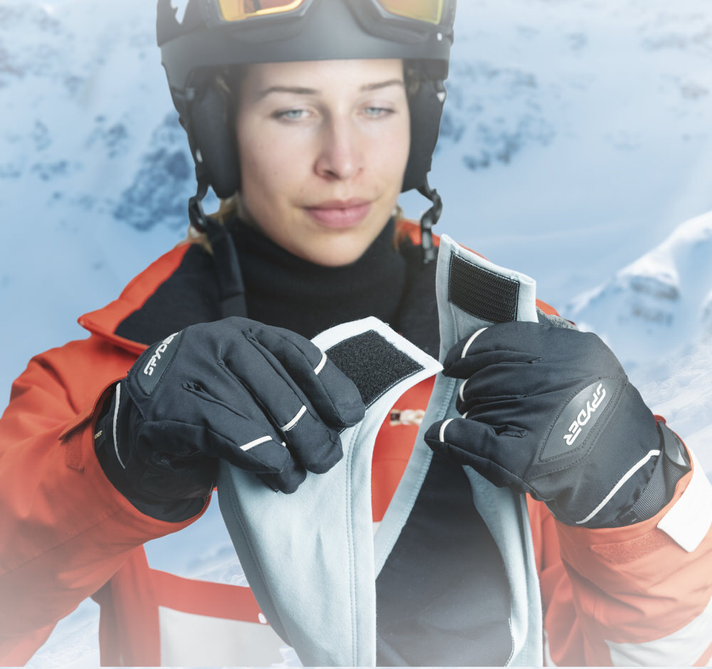 Frau mit Helm und Handschuhen schliesst hellblaue HeiQ Filter Bandana goSnow mit Klettverschluss vor schneebedeckter Bergkulisse.