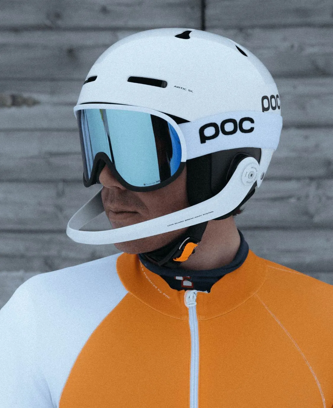 Persona con casco da sci e occhiali da sci POC Retina Race con lenti blu davanti a una parete di legno grigia.