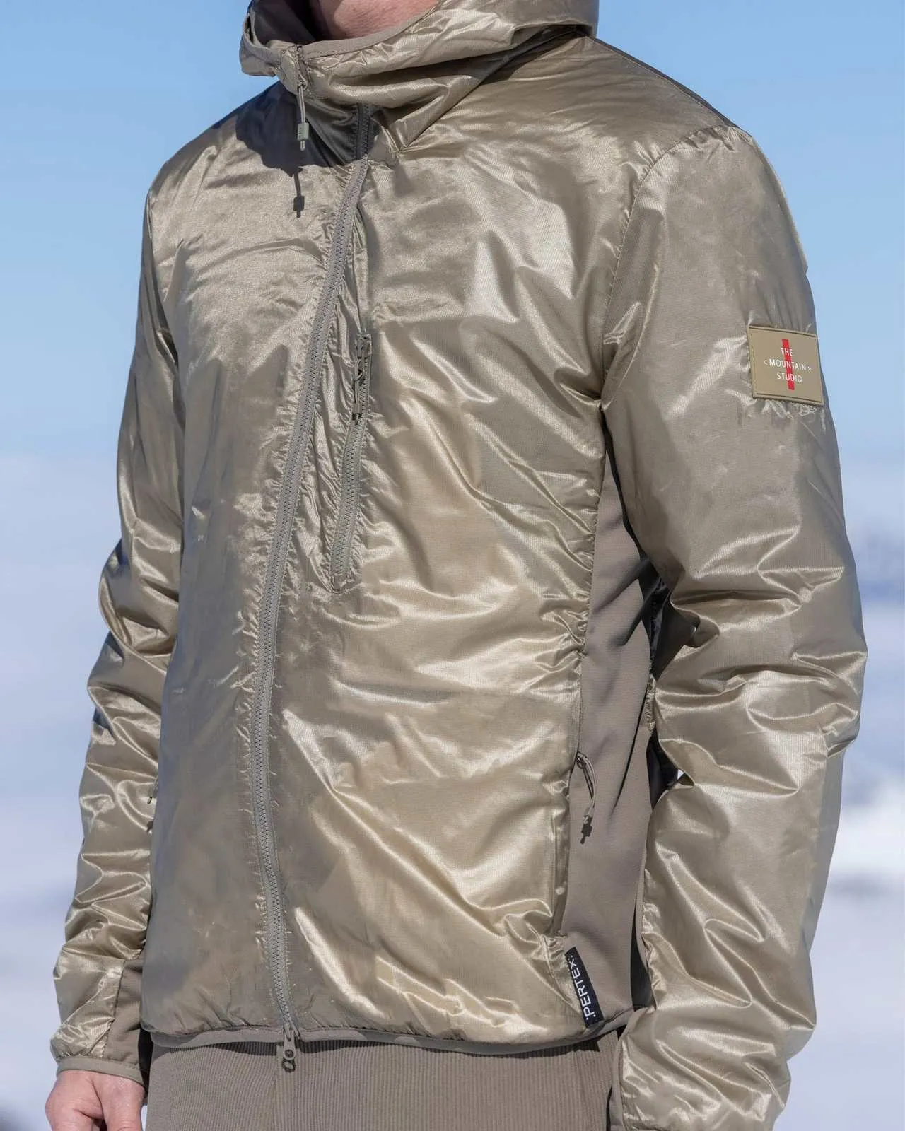 Man in shiny, beige jacket with hood against a snowy background. Details: zippers, sleeve patch, reflective material.