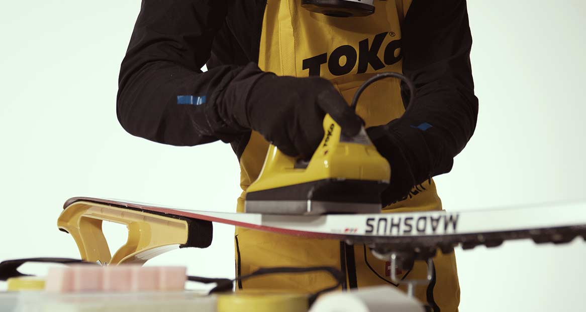 Eine Person mit Handschuhen wachst einen Ski mit einem gelben Bügeleisen über einem Wachsblock.