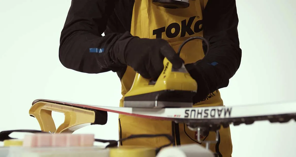 Eine Person mit Handschuhen wachst einen Ski mit einem gelben Bügeleisen über einem Wachsblock.