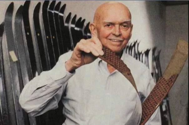 Un uomo sorridente e di carnagione chiara tiene in mano degli sci con un motivo a rete davanti a uno scaffale pieno di sci. Indossa una camicia chiara.