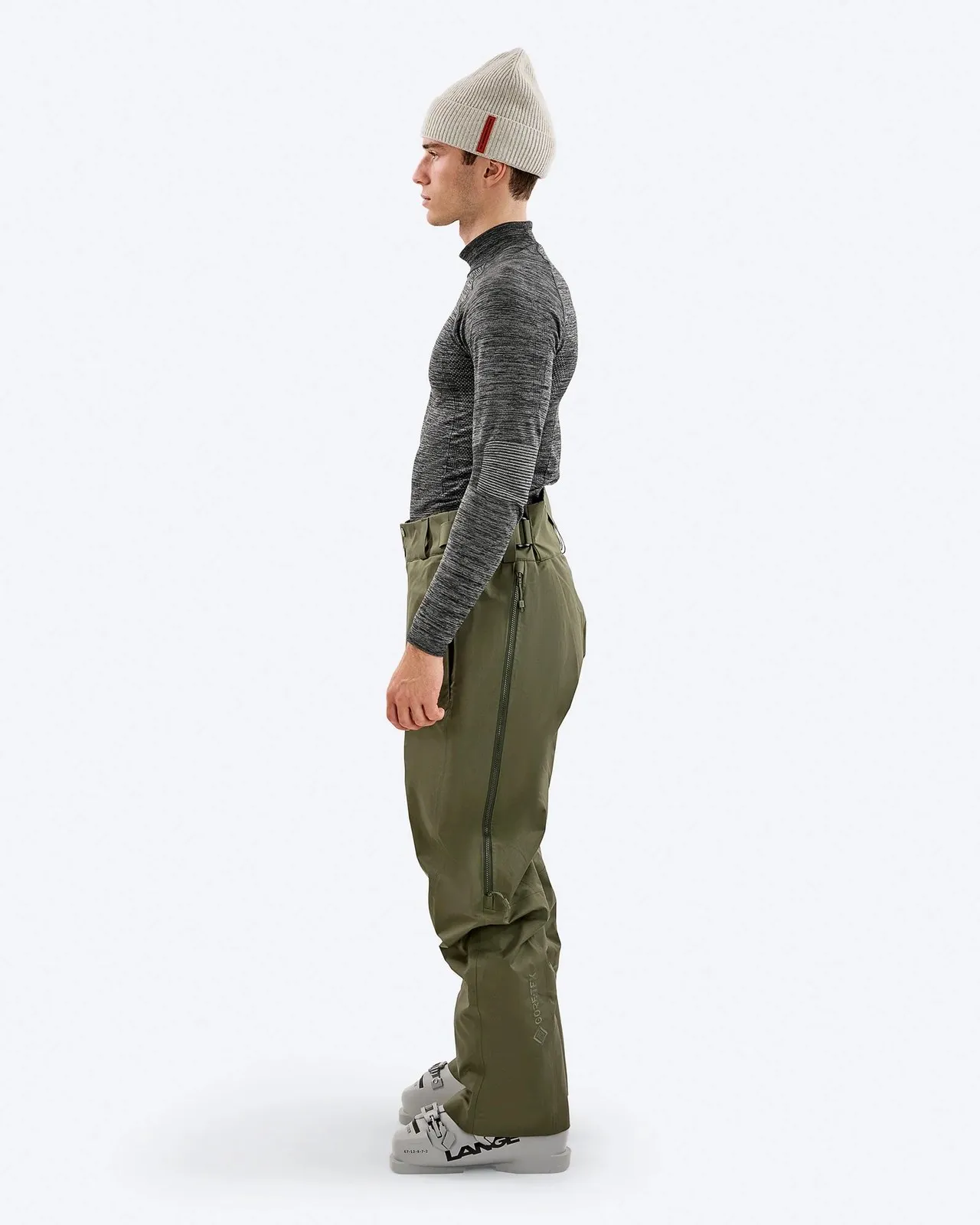 A man in ski clothing stands sideways in front of a white background. He is wearing a gray cap, a long-sleeved shirt and olive green ski pants and ski boots.