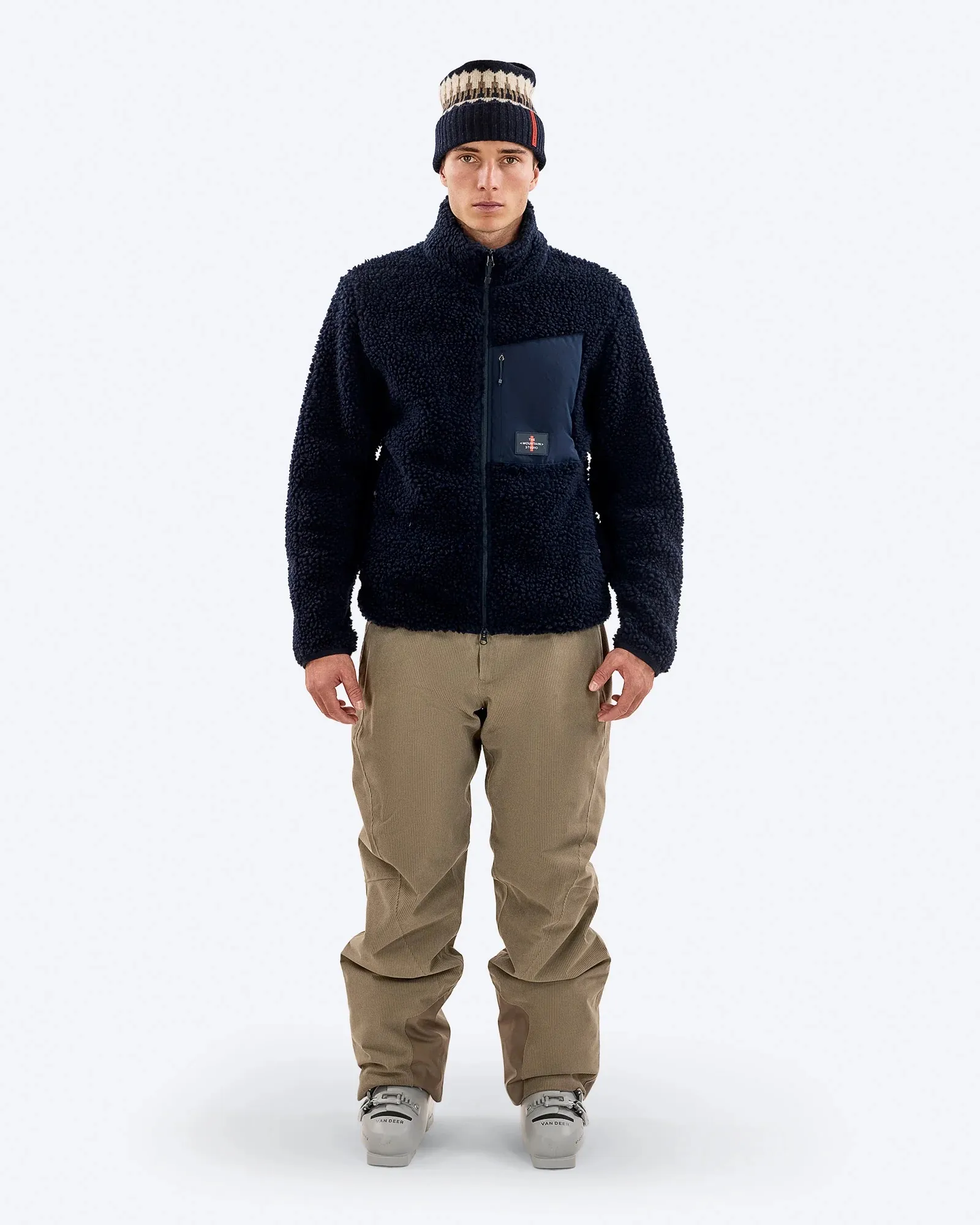Uomo con giacca di pile blu, pantaloni di velluto a coste e berretto, in piedi davanti a uno sfondo bianco. Sono visibili gli scarponi da sci.