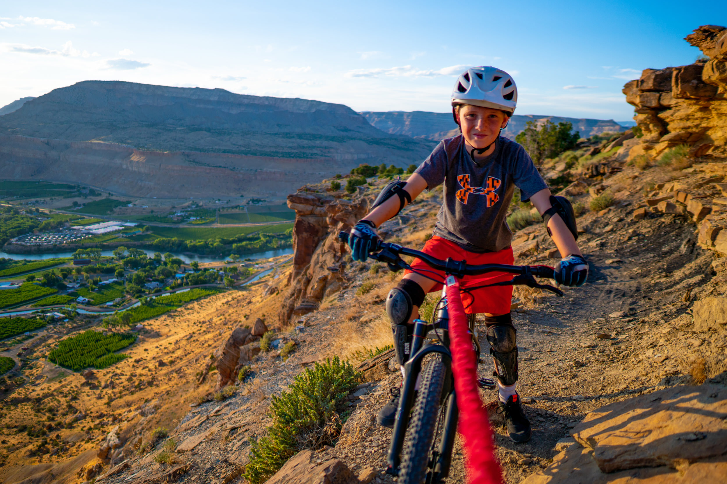 Kind mit Helm und Schutzausrüstung auf einem Mountainbike mit Tow-Whee Abschleppseil vor einer Berglandschaft.
