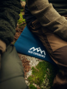 Beheiztes Sitzkissen im Freien Person sitzt auf blauem, beheiztem Sitzkissen mit Logo im Freien auf moosbewachsenem Untergrund.