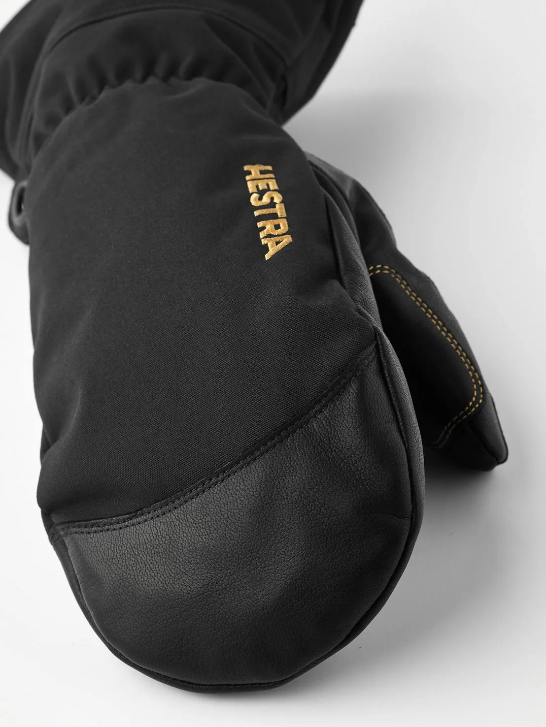 Close-up of a black Hestra mitten made of leather and fabric, with the gold-colored Hestra logo on the back of the hand.