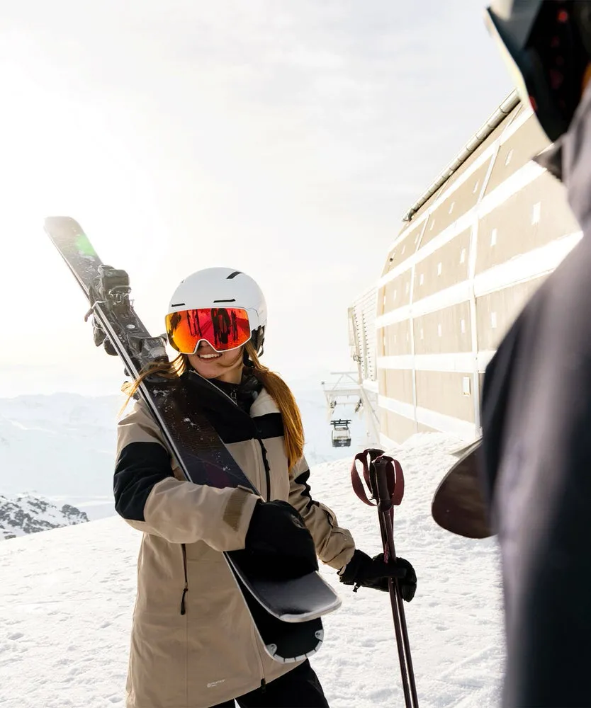 Skier with white helmet (Driver Prime Sigma Photo Mips) and skis on her shoulder in the snow. Sunlight and mountain panorama in the background.