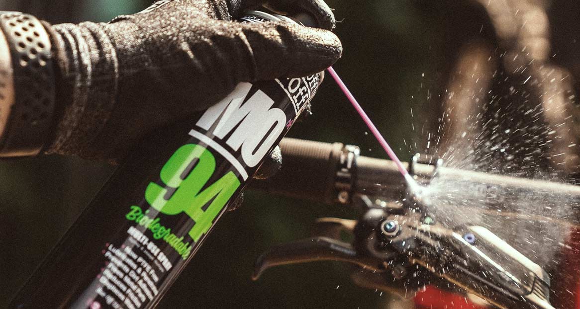 Une main portant un gant noir vaporise du détergent sur un vélo à l'aide d'une boîte de "MO-94", ce qui provoque des éclaboussures.
