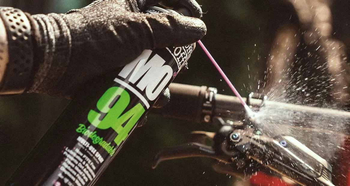 Eine Hand in einem schwarzen Handschuh sprüht mit einer Dose "MO-94" Reinigungsmittel auf ein Fahrrad, wodurch Spritzer entstehen.
