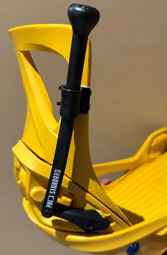 Close-up of a bright yellow snowboard binding against a light brown background. A black adjustment lever labeled “Pro Standard” runs vertically and is attached to the highback for precise adjustment.