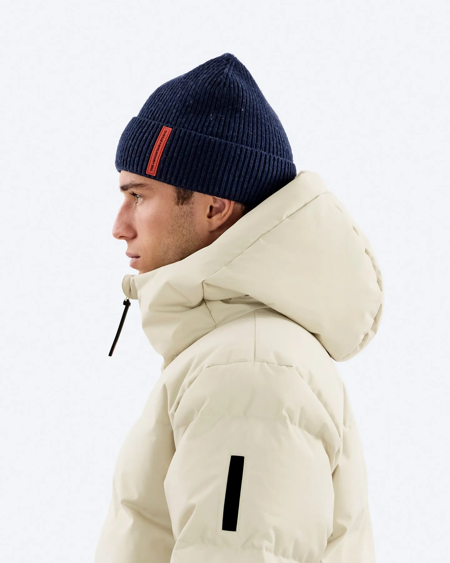 Profilo di un uomo con berretto blu scuro e piumino beige davanti a uno sfondo neutro.