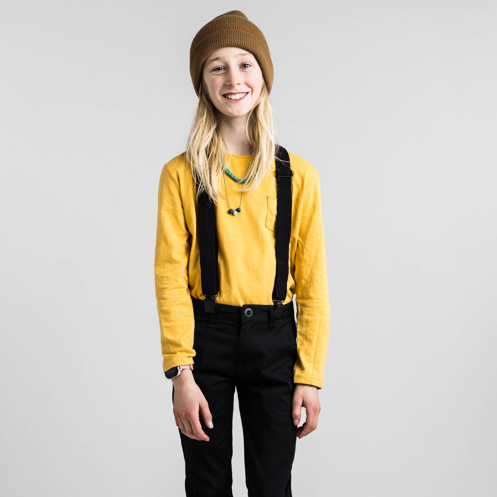 Jeune fille souriante avec une casquette marron, un haut jaune, un pantalon noir et des bretelles sur un fond gris.