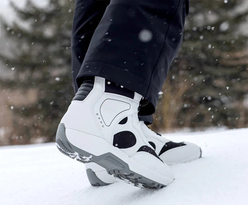Weiße Dahu Skischuhe Eine Person trägt weiße Dahu Skischuhe mit schwarzen Akzenten im Schnee. Im Hintergrund sind Bäume zu sehen.