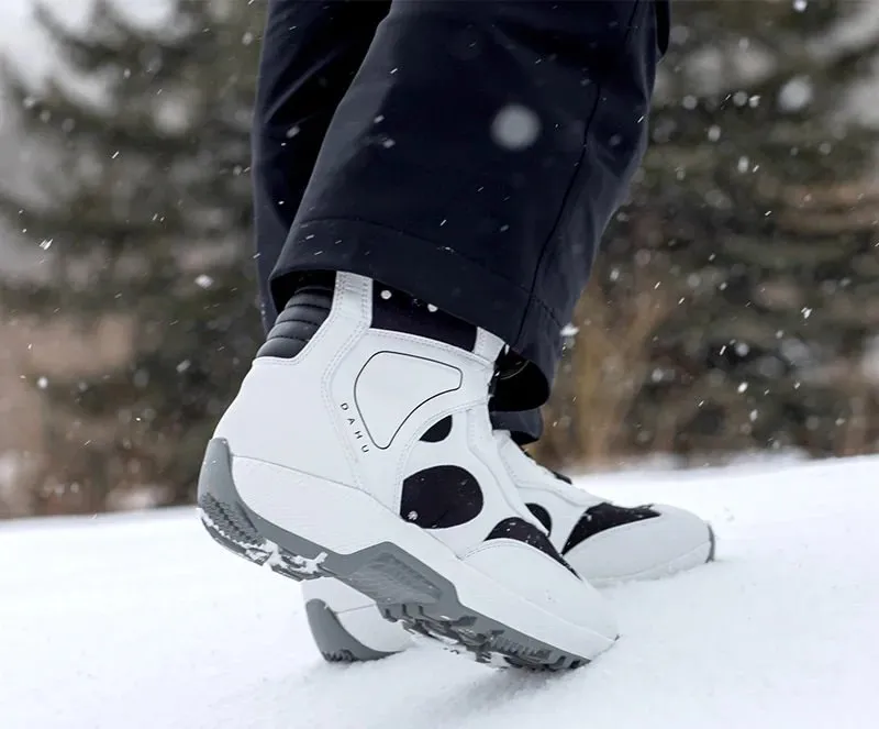 Une personne porte des chaussures de ski blanches Dahu avec des accents noirs dans la neige. Des arbres sont visibles en arrière-plan.