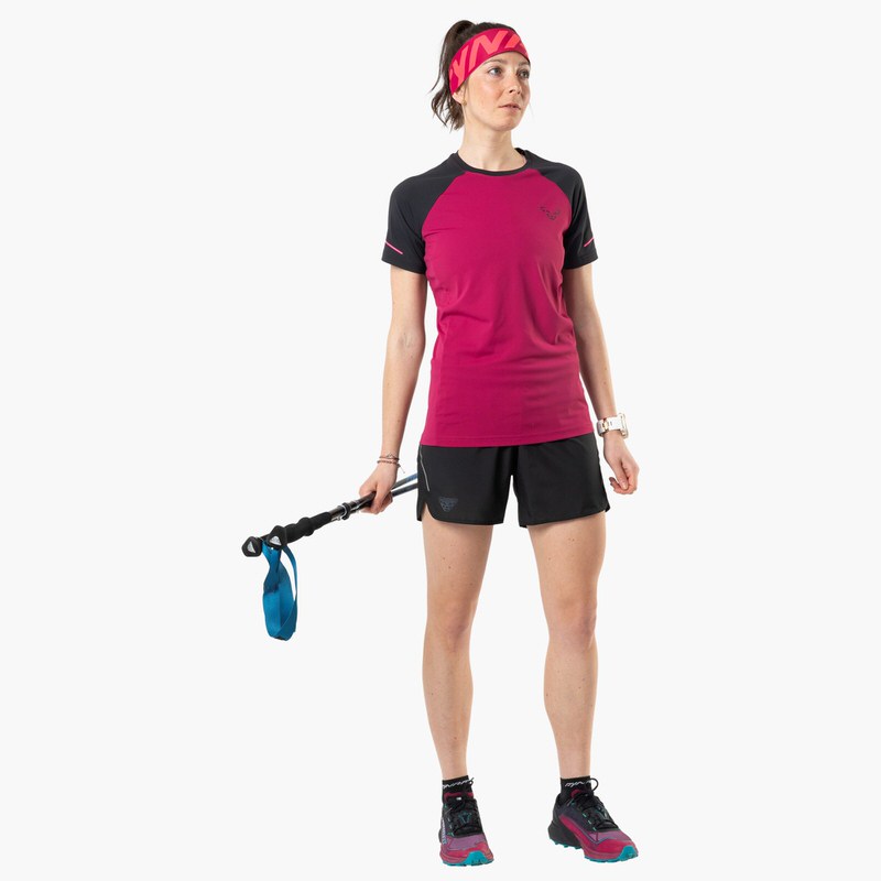 Frau im Alpine Pro Shirt in Black Out Beet Red mit Trekkingstöcken, Shorts und Stirnband.