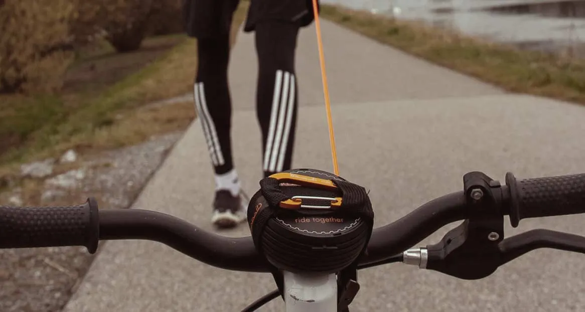 Fahrradlenker mit montiertem Abschleppseil; im Hintergrund eine Person in Sportkleidung auf einem Weg.