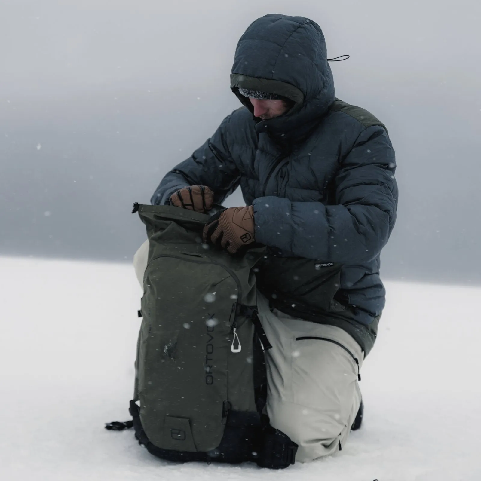 Un uomo inginocchiato nella neve, indossa un piumino e apre uno zaino verde oliva. Nevica leggermente.