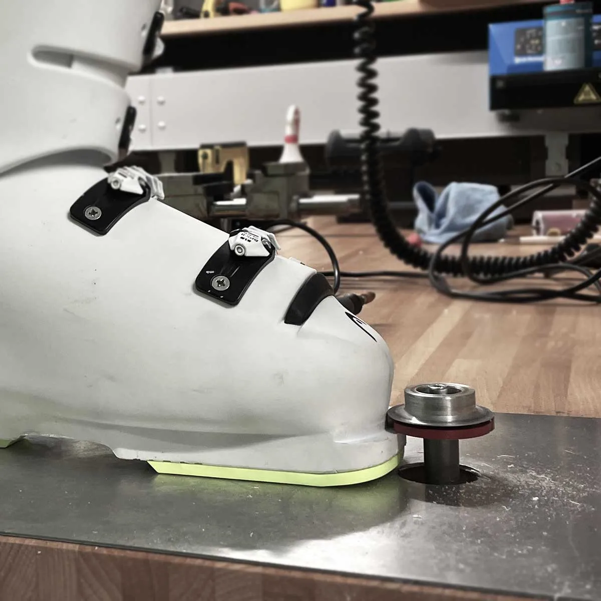 Une chaussure de ski blanche est posée sur une plaque métallique, avec un dispositif métallique sous la chaussure. L'arrière-plan de l'atelier est flou.