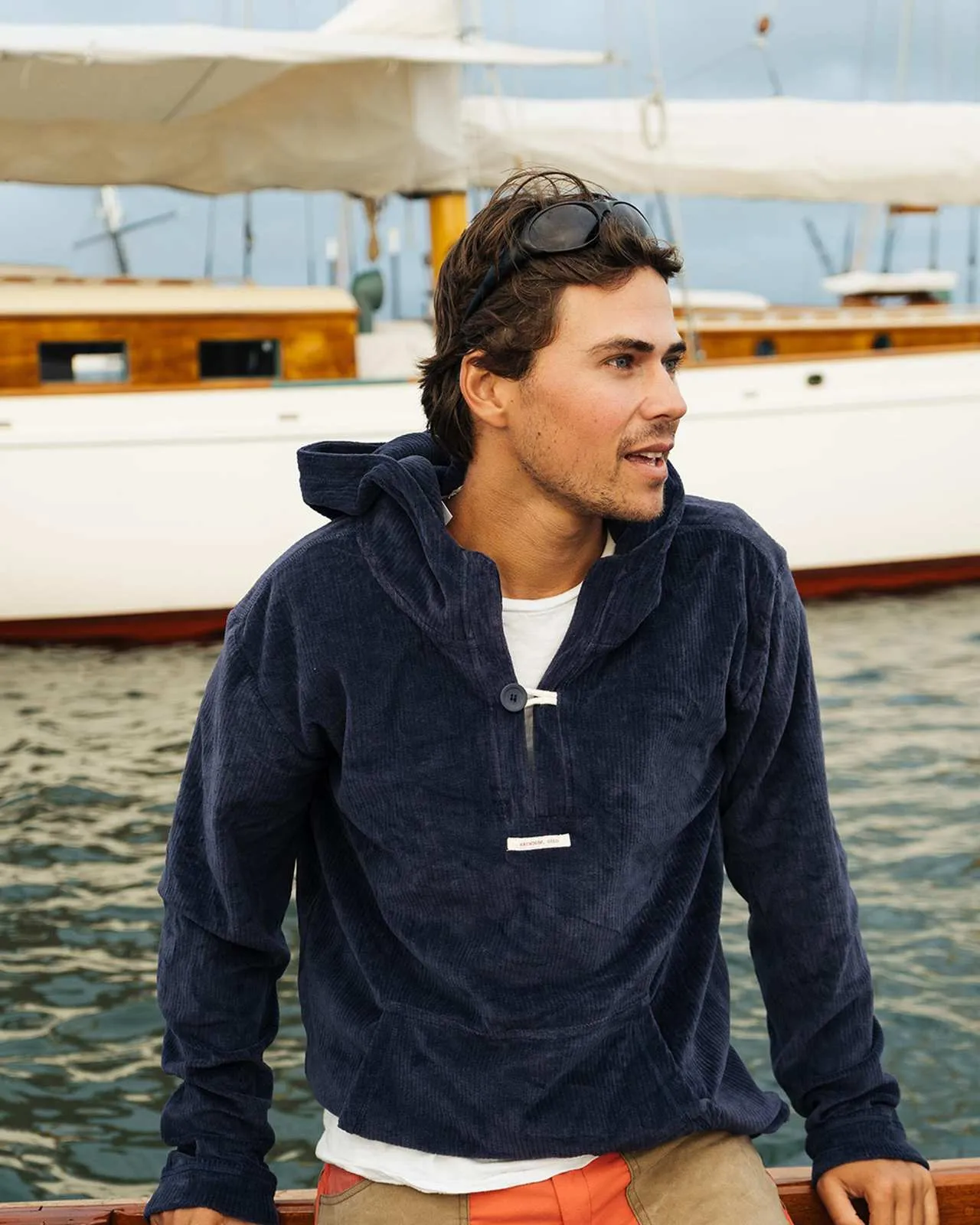 A man in a blue corduroy hoodie is leaning against a yacht railing. He wears sunglasses on his head and looks to the right.
