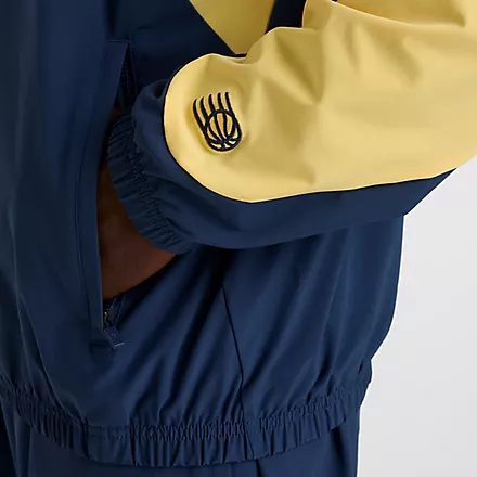 New Balance Track Jacket Detailaufnahme der New Balance In Game Woven Track Jacket in Navy/Gelb. Sichtbar: Ärmel-Logo, Seitentasche mit Reißverschluss und elastischer Saum.