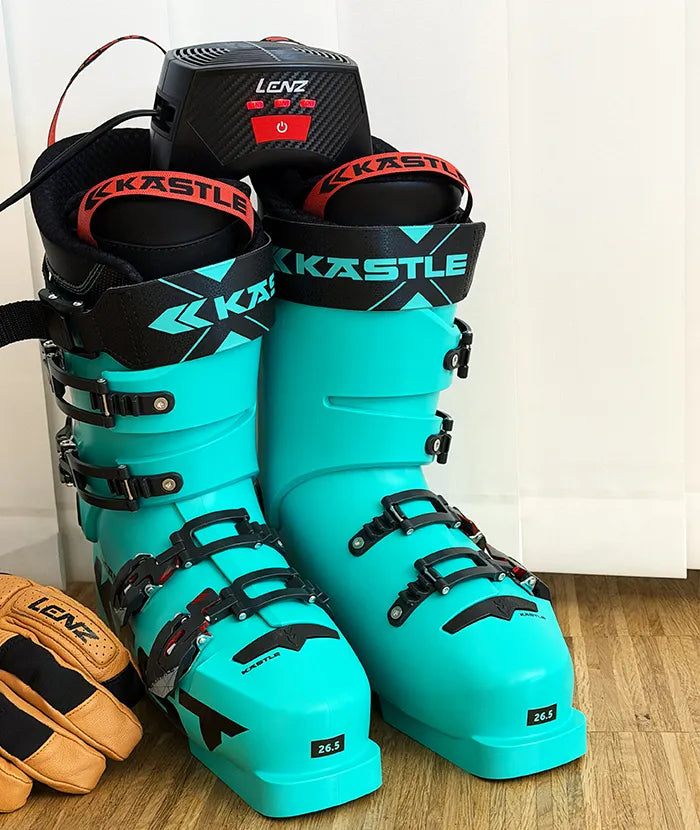 A pair of turquoise ski boots from the brand Kästle stand on a wooden floor, with a black Lenz heater on top and a pair of Lenz gloves next to them.