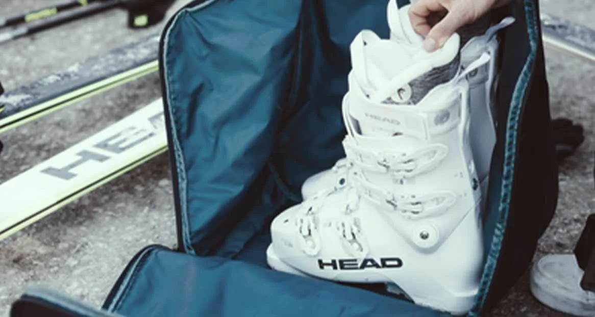 An open ski boot bag with a white HEAD ski boot, skis and ski poles lying next to it.