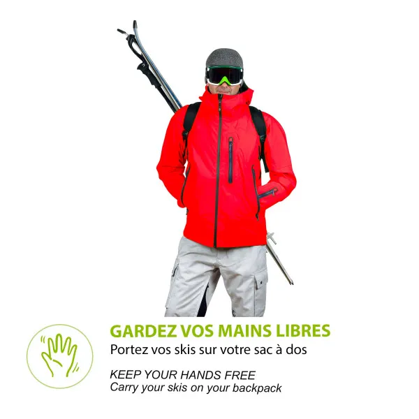 Man in red jacket, gray trousers, cap and ski goggles, carrying skis with ski pack on backpack.