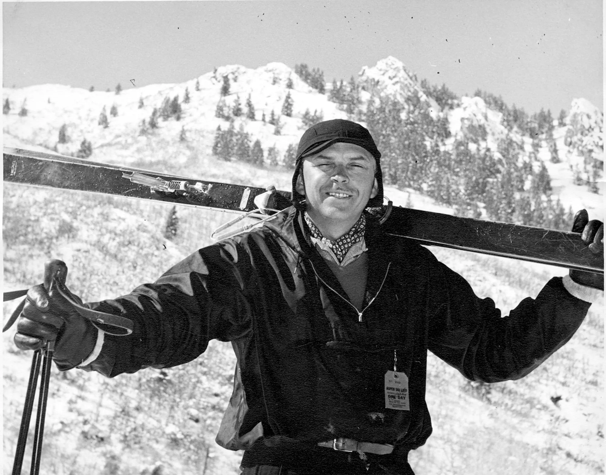 Ritratto in bianco e nero di un uomo sorridente con gli sci sulle spalle davanti a montagne innevate.