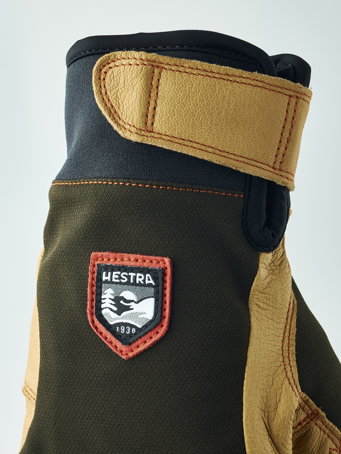 Detailaufnahme eines Handschuhs mit braunem Obermaterial, Lederdetails und "Hestra 1936" Logo. Der Handschuh hat eine Stulpe mit Klettverschluss.