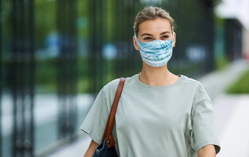 Femme portant un masque à filtre bleu clair Buff à l'extérieur, portant un T-shirt clair et un sac.