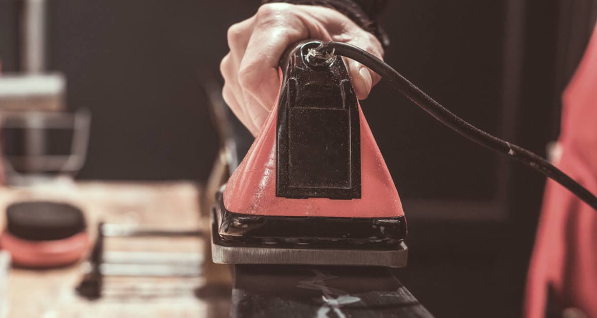 Eine Hand hält ein Wachseisen mit roter und schwarzer Oberfläche, ein Kabel hängt herunter. Im Hintergrund sind unscharfe Gegenstände.
