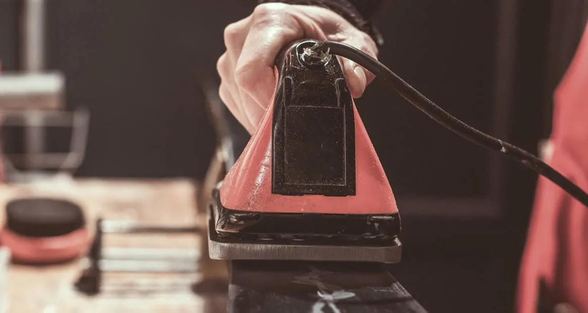 Eine Hand hält ein Wachseisen mit roter und schwarzer Oberfläche, ein Kabel hängt herunter. Im Hintergrund sind unscharfe Gegenstände.