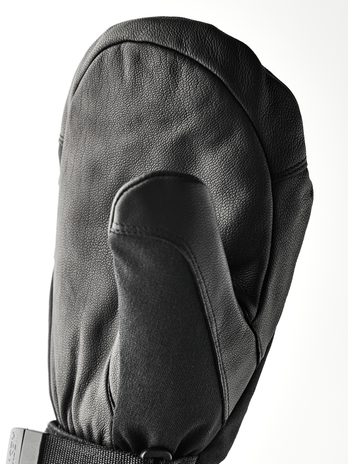 Black leather glove A black, leather mitten glove lies on a white surface. The thumb is pointing forwards and the back of the hand is curved.