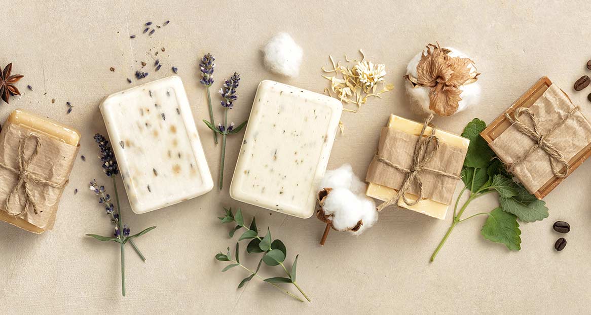 Handseifen mit Lavendel Flach liegende Handseifen, teils verpackt mit Papier und Bindfaden, Lavendelblüten, Baumwolle und Kaffeebohnen auf beigem Grund.