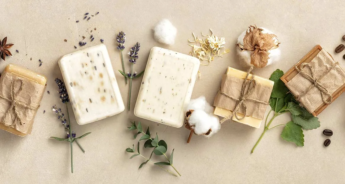 Flach liegende Handseifen, teils verpackt mit Papier und Bindfaden, Lavendelblüten, Baumwolle und Kaffeebohnen auf beigem Grund.