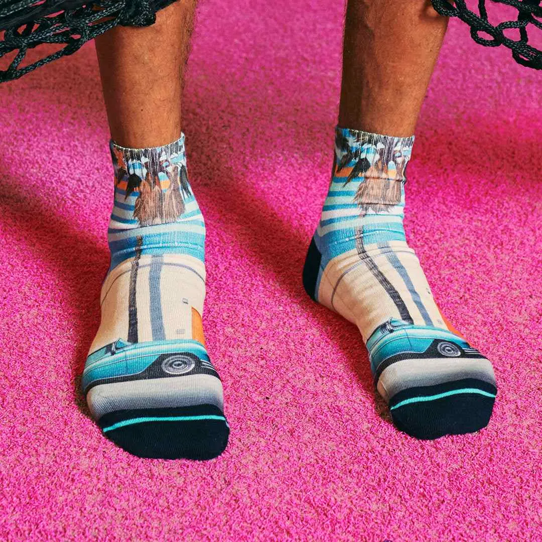 Close-up of two feet in quarter socks with Miami motif on pink carpet. Palm trees, beach and car are depicted.