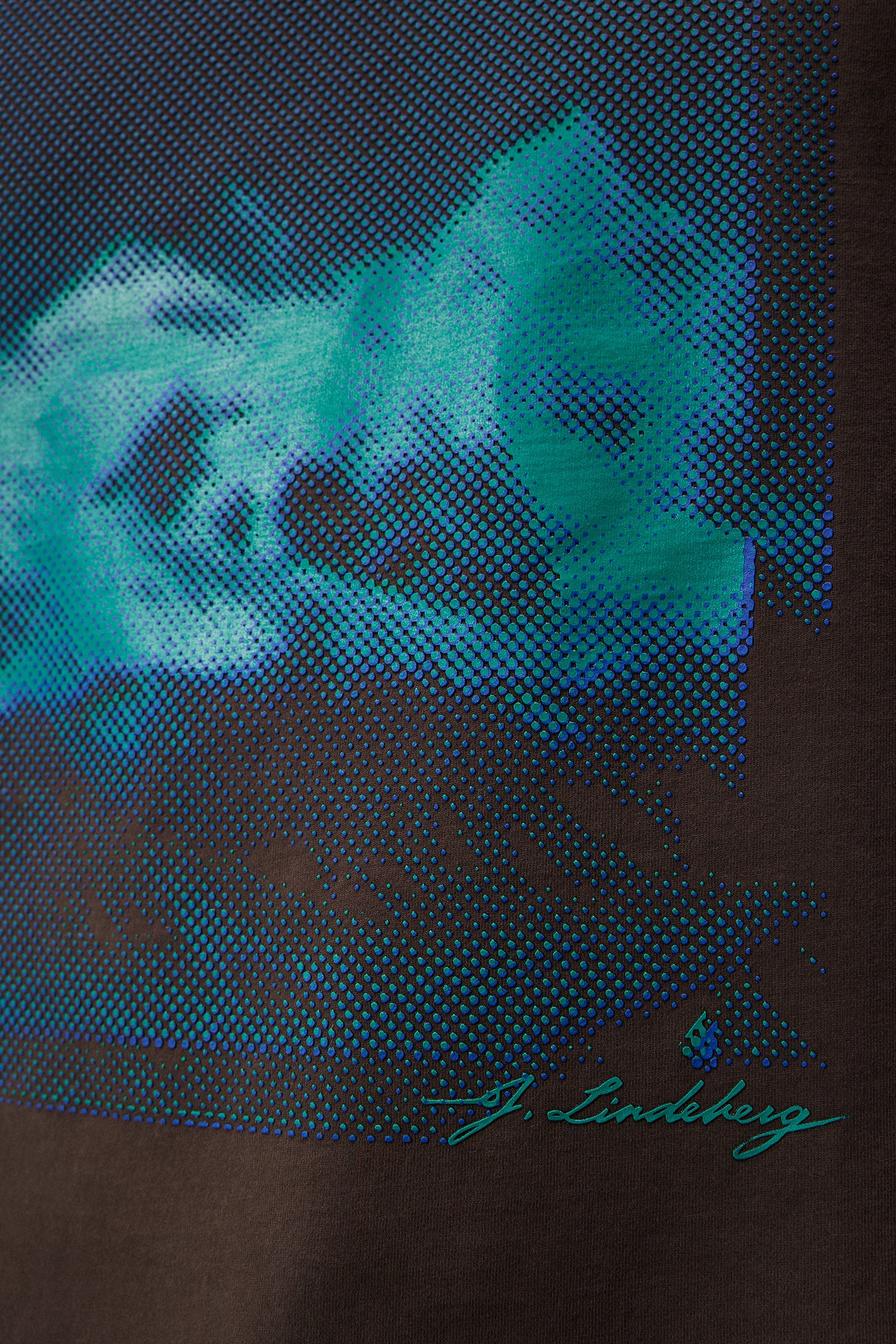 Detailaufnahme des Walsh Boxy Print Tee Delicioso mit blau-grünem, saisonalem Print und J. Lindeberg Schriftzug auf braunem Stoff.