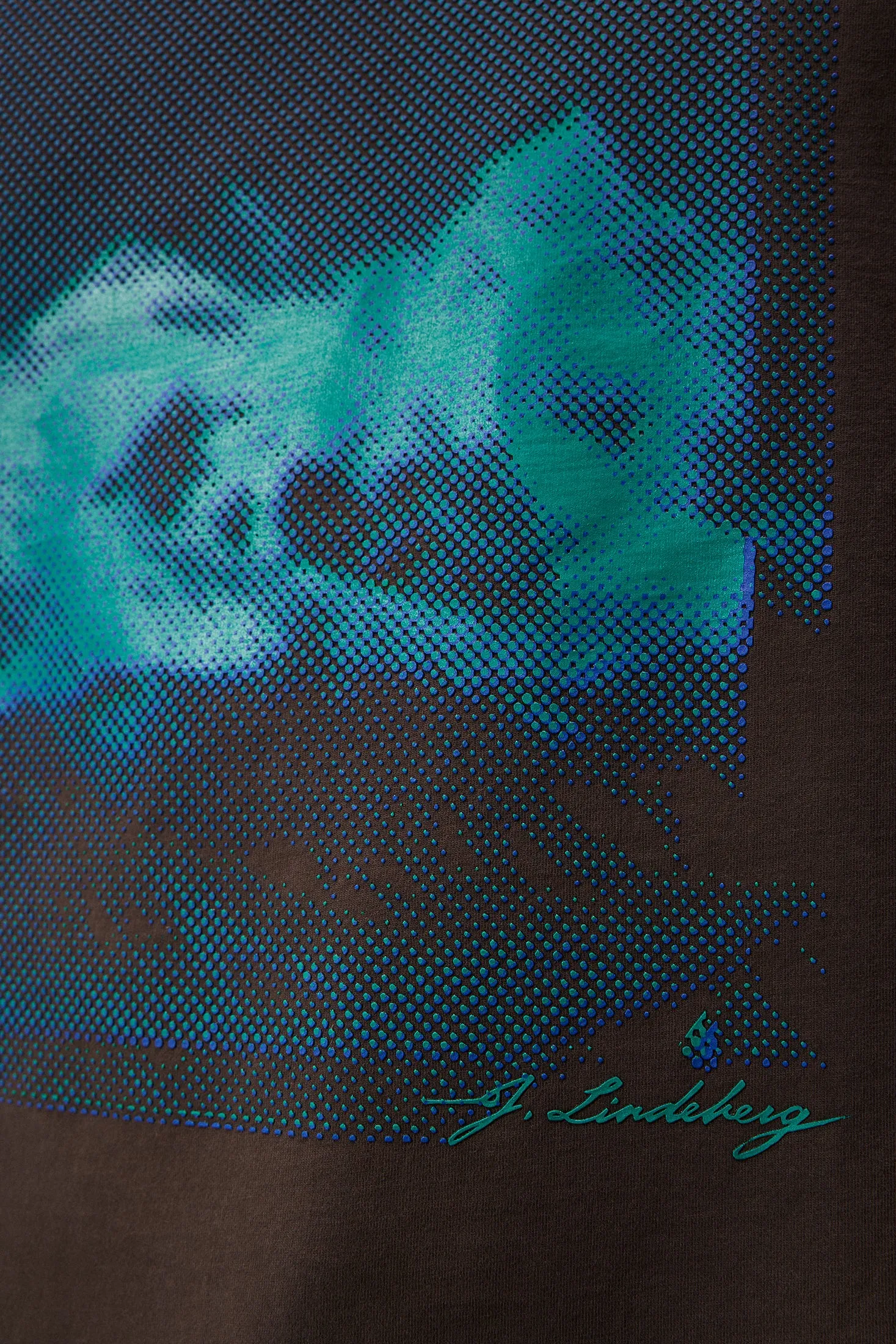 Detailaufnahme des Walsh Boxy Print Tee Delicioso mit blau-grünem, saisonalem Print und J. Lindeberg Schriftzug auf braunem Stoff.