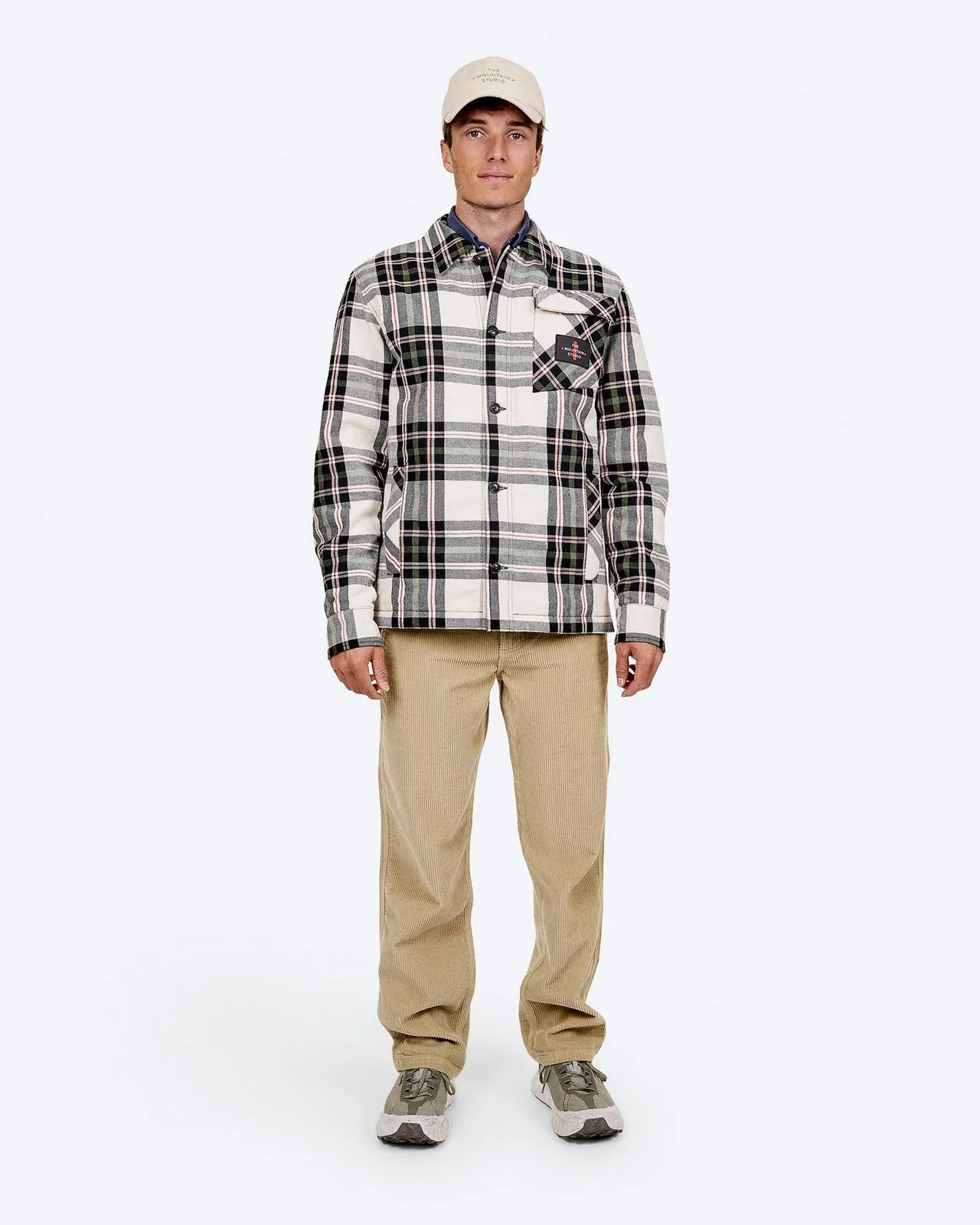 Ein Mann in Cordhose, karierter Jacke und Mütze steht vor einem weißen Hintergrund.