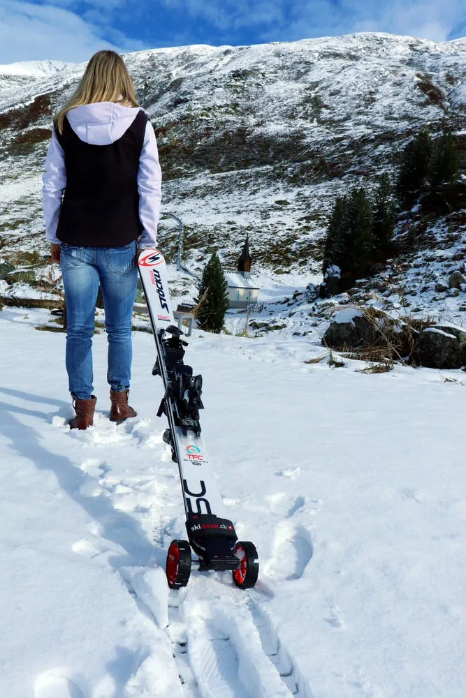 Woman with skis on snow, equipped with red Skimover Traveller Pro with wheels. A mountain and blue sky are visible in the background.
